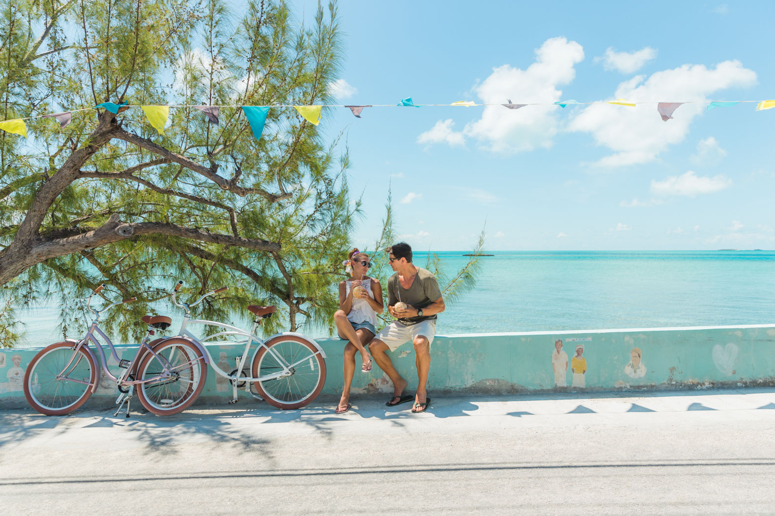 A couple enjoys a leisurely afternoon on a wall next to a tree, their bicycles parked nearby.