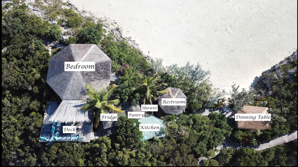 Aerial view of a beachside cabin on a private island in the Exuma Cays, labeled with Bedroom, Kitchen, Restroom, Dining Table, Shower, Pantry, Fridge, and Deck. The cabin is surrounded by lush trees and a pristine sandy beach.