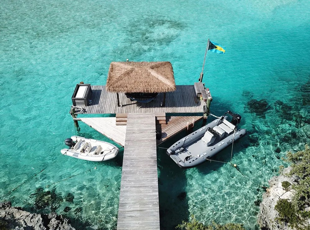 A wooden dock with a thatched roof hut extends into the clear turquoise waters of the Exuma Cays, with two boats moored on either side and a flag flying at the end.