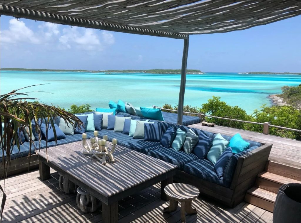 A shaded outdoor seating area with blue cushions overlooks the turquoise Exuma Cays and a distant green island under a clear blue sky. Private Island Vacation Package Bahamas Island Travel