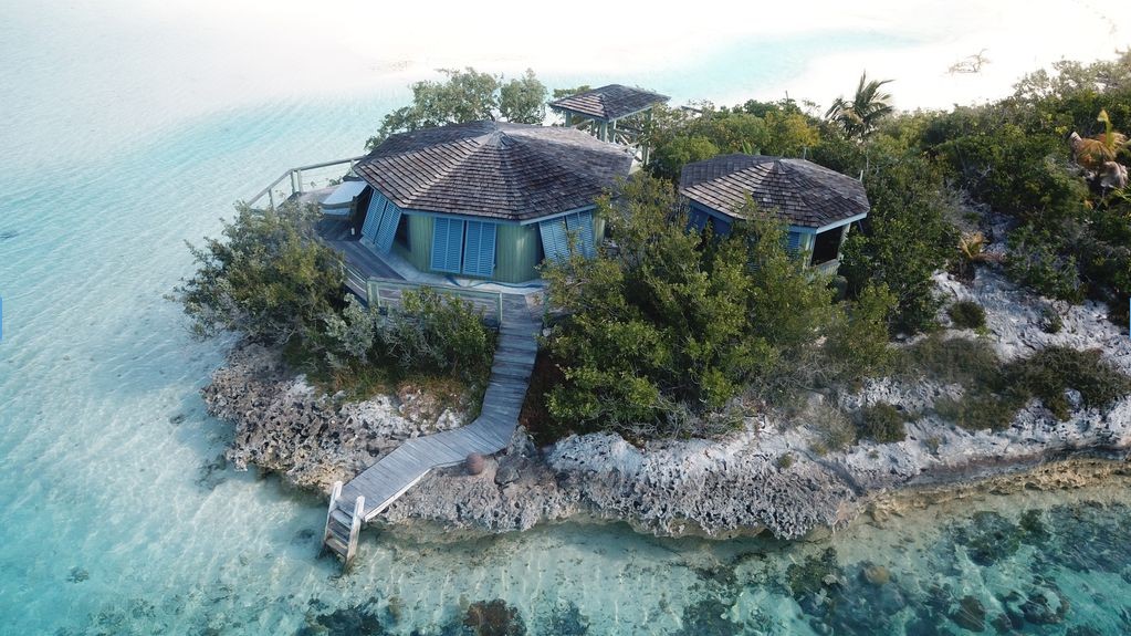 A small, round building with a wooden walkway sits on a rocky, bush-covered private island in the Exuma Cays, surrounded by clear blue water. Private island rental vacation pagckage Bahamas Island Travel