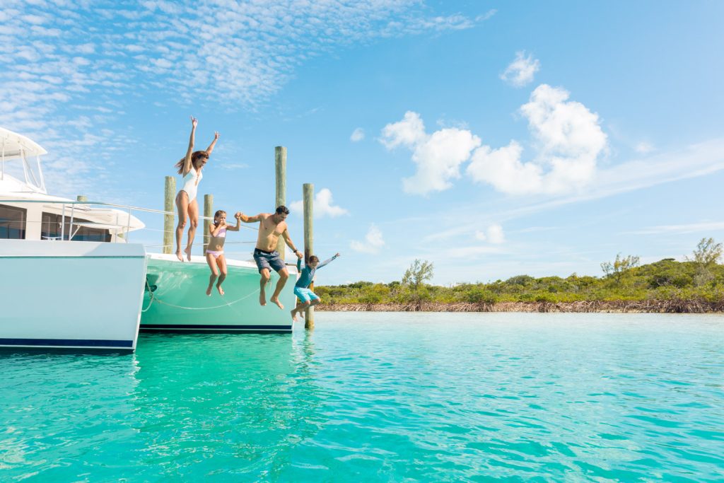 A group of people jump off a boat into clear blue water, with a green shoreline and blue sky in the background.