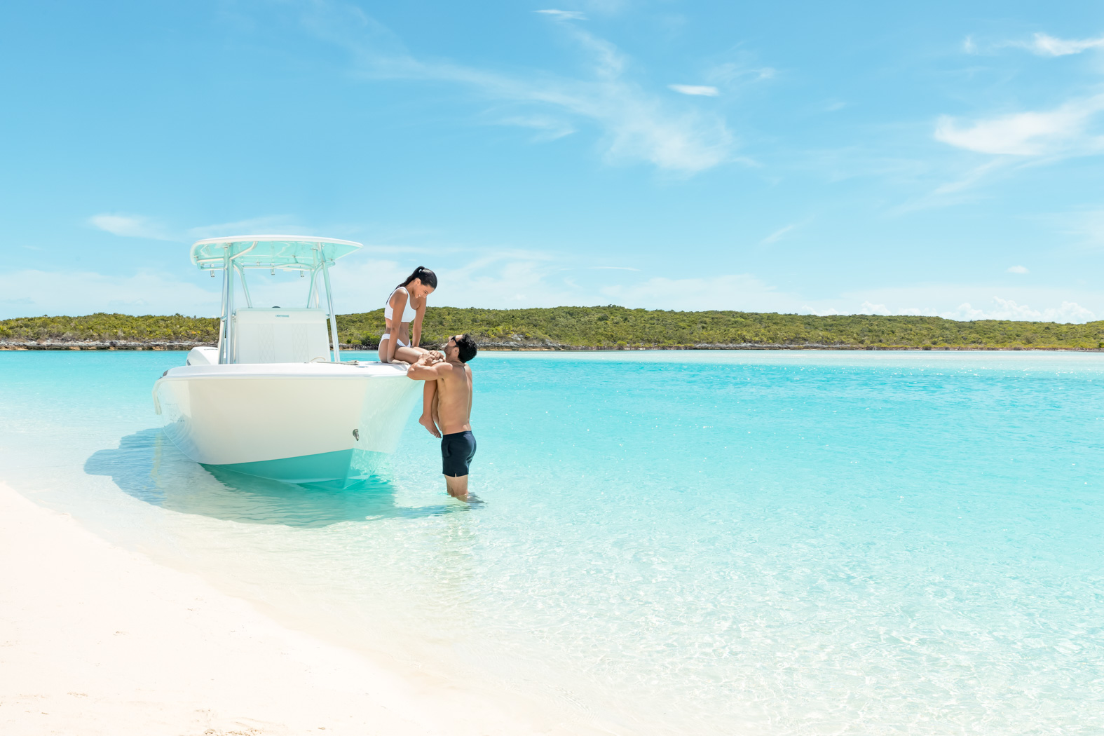 A man in swim trunks helps a woman in a swimsuit onto a white boat in clear shallow water near a sandy beach with a greenery-covered shoreline under a sunny, blue sky.