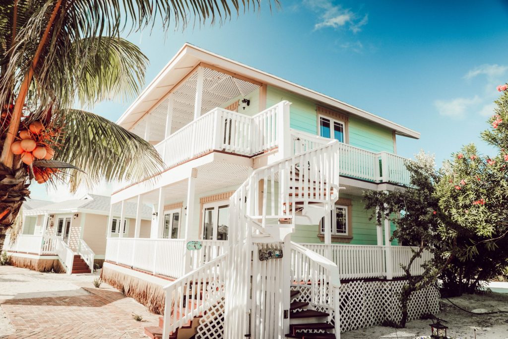 A pastel-colored two-story house with a wraparound porch and white spiral staircase, surrounded by tropical plants under a clear blue sky.