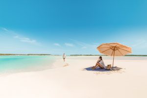 A person sits under a beach umbrella on a white sandy shore with turquoise water, while another person walks along the waterline. The sky is clear and blue.