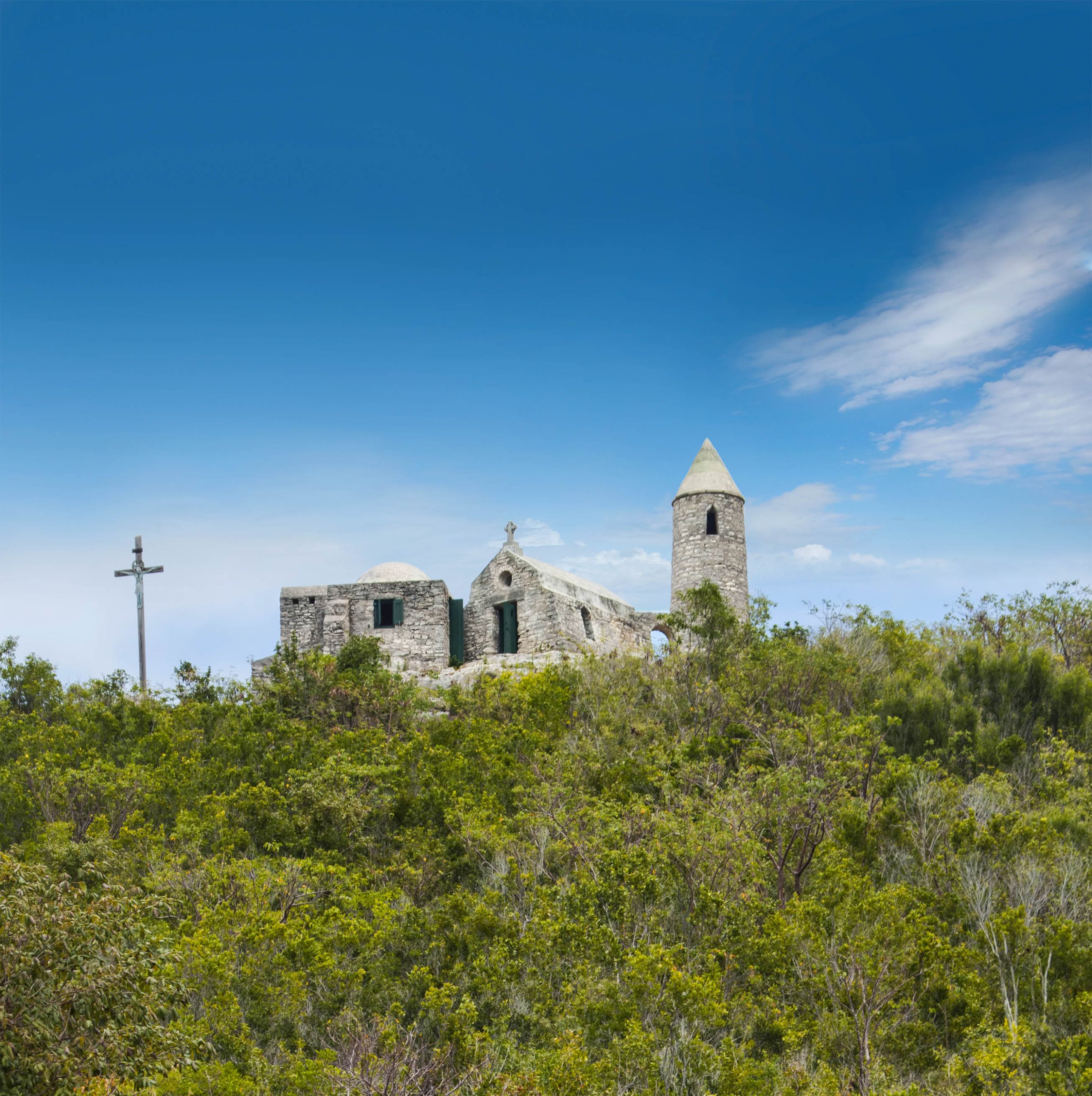 A stone church with a steeple sits atop a green hill under a clear blue sky. A tall cross stands to the left of the church.