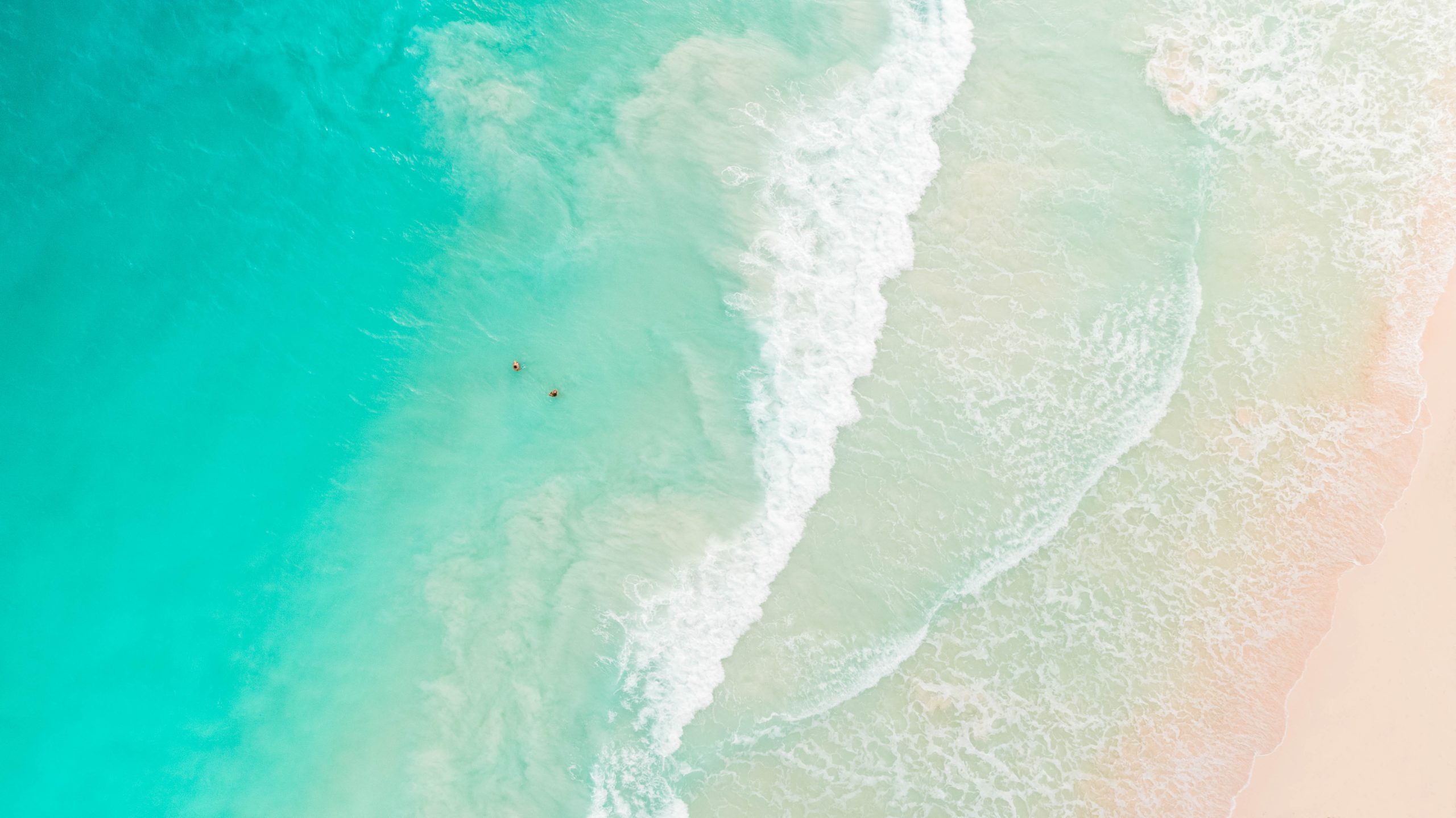 Aerial view of a turquoise ocean meeting a sandy beach, with two people swimming near the shore. Waves are gently breaking onto the beach.