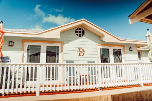 A light green house with white trim and a deck with white railing under a blue sky. There are two lounge chairs on the deck and an emblem with "D" and "L" on the house. Vacation Rental on Staniel Cay