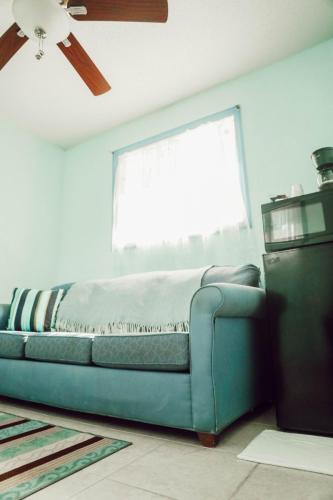Vacation Rental on Staniel Cay A room with light blue walls features a blue sofa with a light blanket, a striped cushion, a window with sheer curtains, a ceiling fan, a small refrigerator, and a coffee maker on top.