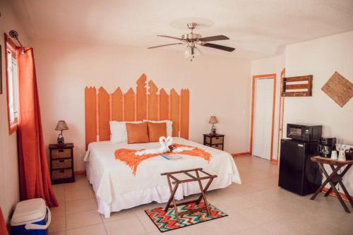 A cozy hotel room with a king-sized bed adorned with orange accents and a towel sculpture. The room includes a ceiling fan, small fridge, microwave, side tables, and a small dining area. Vacation Rental on Staniel Cay Named Cat Island