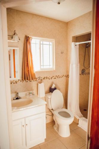 A bathroom with beige tiles features a vanity with a sink, a toilet, and a shower with a glass door. A window with orange curtains is above the toilet. Vacation Rental on Staniel Cay Named Cat Island