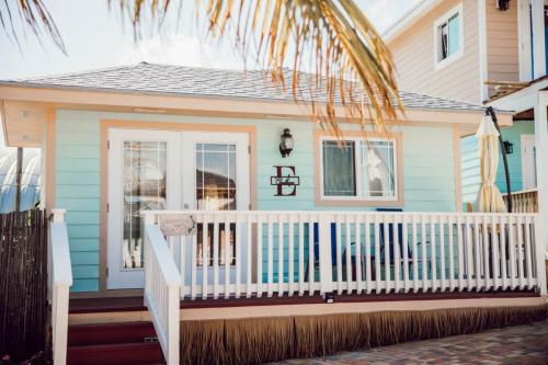 A small, light-blue cottage with white trim, double glass doors, and a porch with a white railing. A palm leaf partially frames the image. The letter "E" is displayed next to the front door.