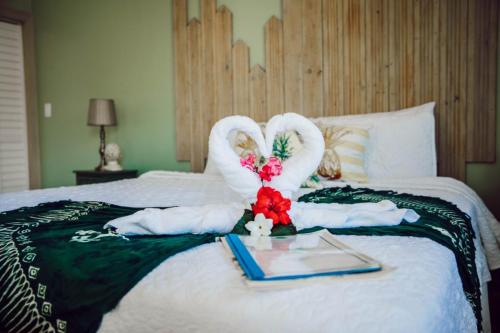 A neatly made bed with white linens, topped with towels folded into swans forming a heart shape. Colorful flowers and a binder are placed in front of the swan towels. A light-green wall and wooden decor are in the background.