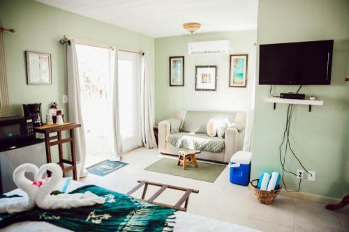 Cozy room with a green theme, featuring a small sofa, wall-mounted TV, coffee maker, mini-fridge, and a made bed. Towels folded into swan shapes are placed on the bed. Sunlight filters through the window.
