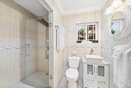A clean, well-lit bathroom in this Staniel Cay vacation rental features a glass-enclosed shower, toilet, and a white sink vanity beneath a round mirror with lights. A window above the sink lets in natural light.