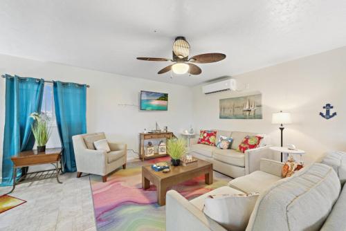 A Staniel Cay vacation rental living room features beige sofas, colorful cushions, a coffee table, two armchairs, a ceiling fan, blue curtains, and nautical-themed decor on white walls. The floor is adorned with a multicolored rug.